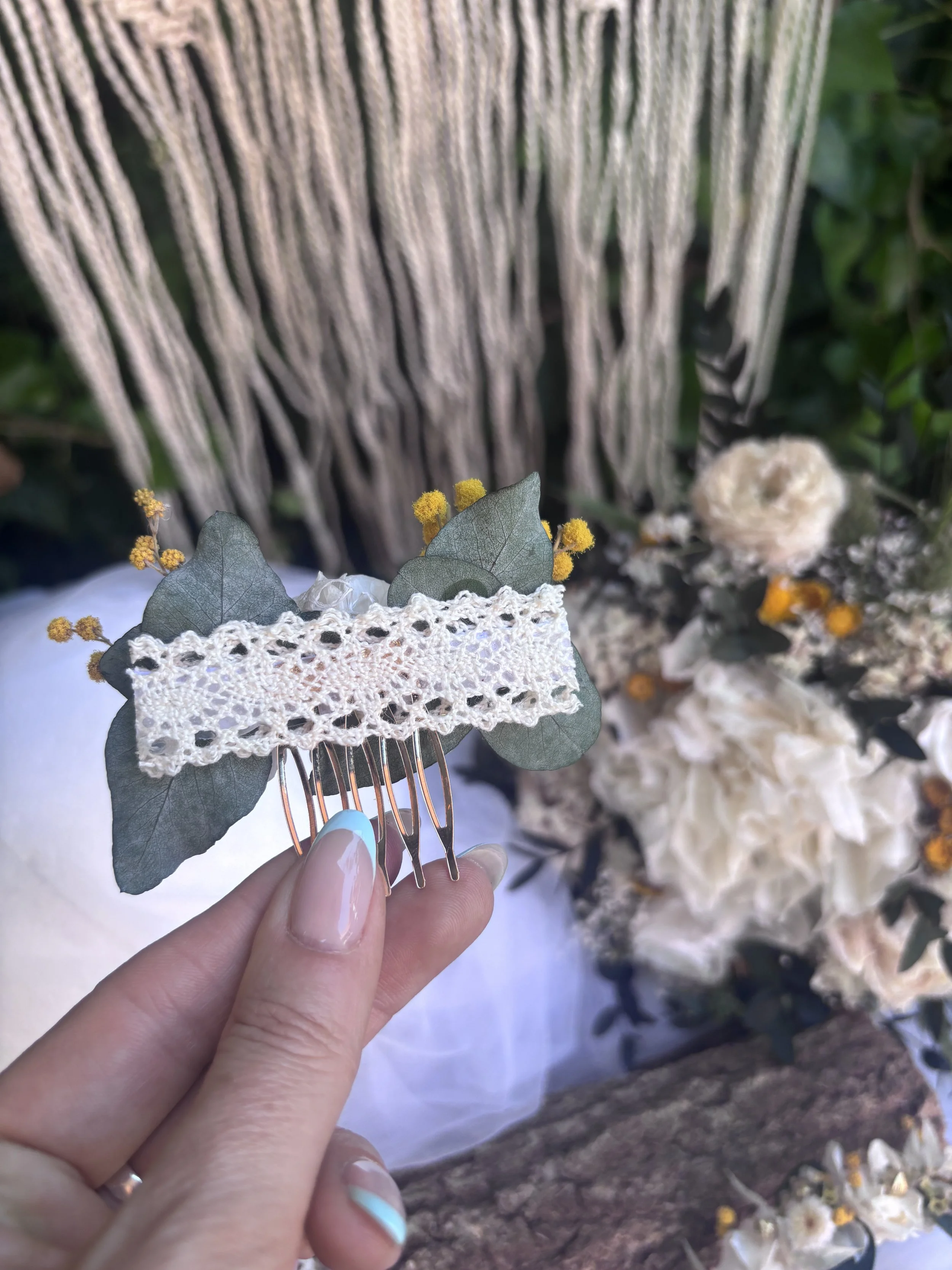 Une main tenant un peigne avec des feuilles vertes, des fleurs blanches et jaunes, et une pièce de dentelle en arrière-plan.