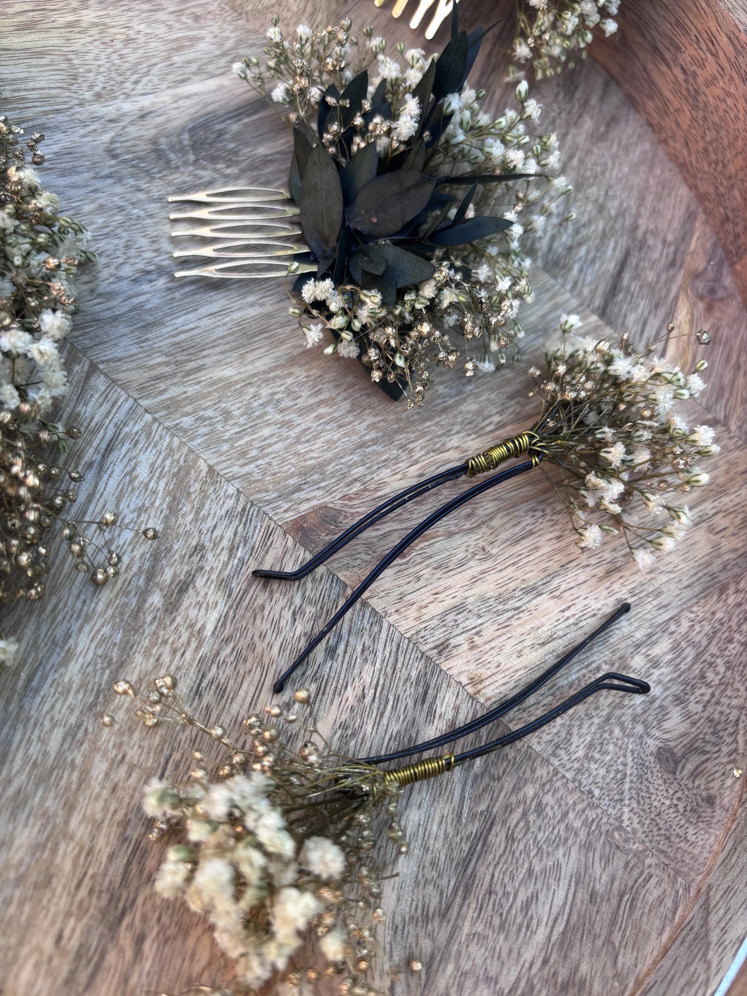 Une couronne de fleurs blanches avec des feuilles vertes foncées et des petites perles dorées, accompagnée d'une bande de fleurs similaires et de plusieurs épingles à cheveux noires avec des détails dorés, posées sur une surface en bois.