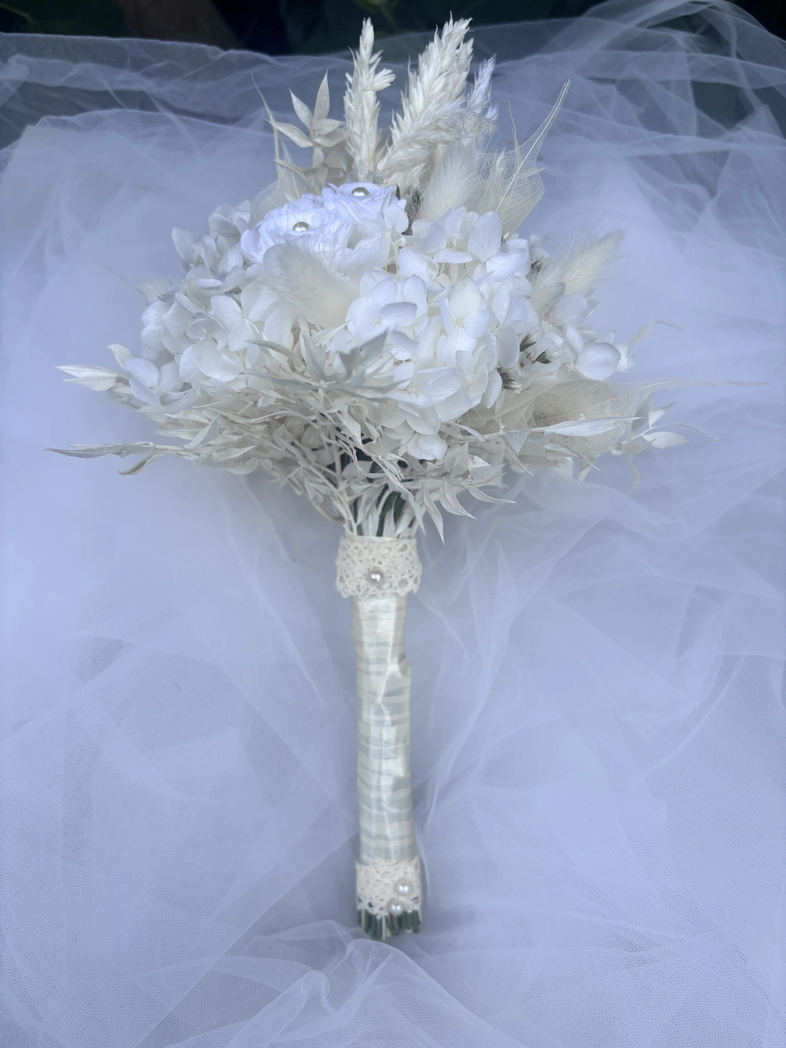 Bouquet de mariée en fleurs séchées et stabilisées blanches, réalisé artisanalement dans un esprit bohème chic et romantique. Cette création florale associe hortensias stabilisés, roses éternelles ornées de perles, arums délicats, feuillages blanchis