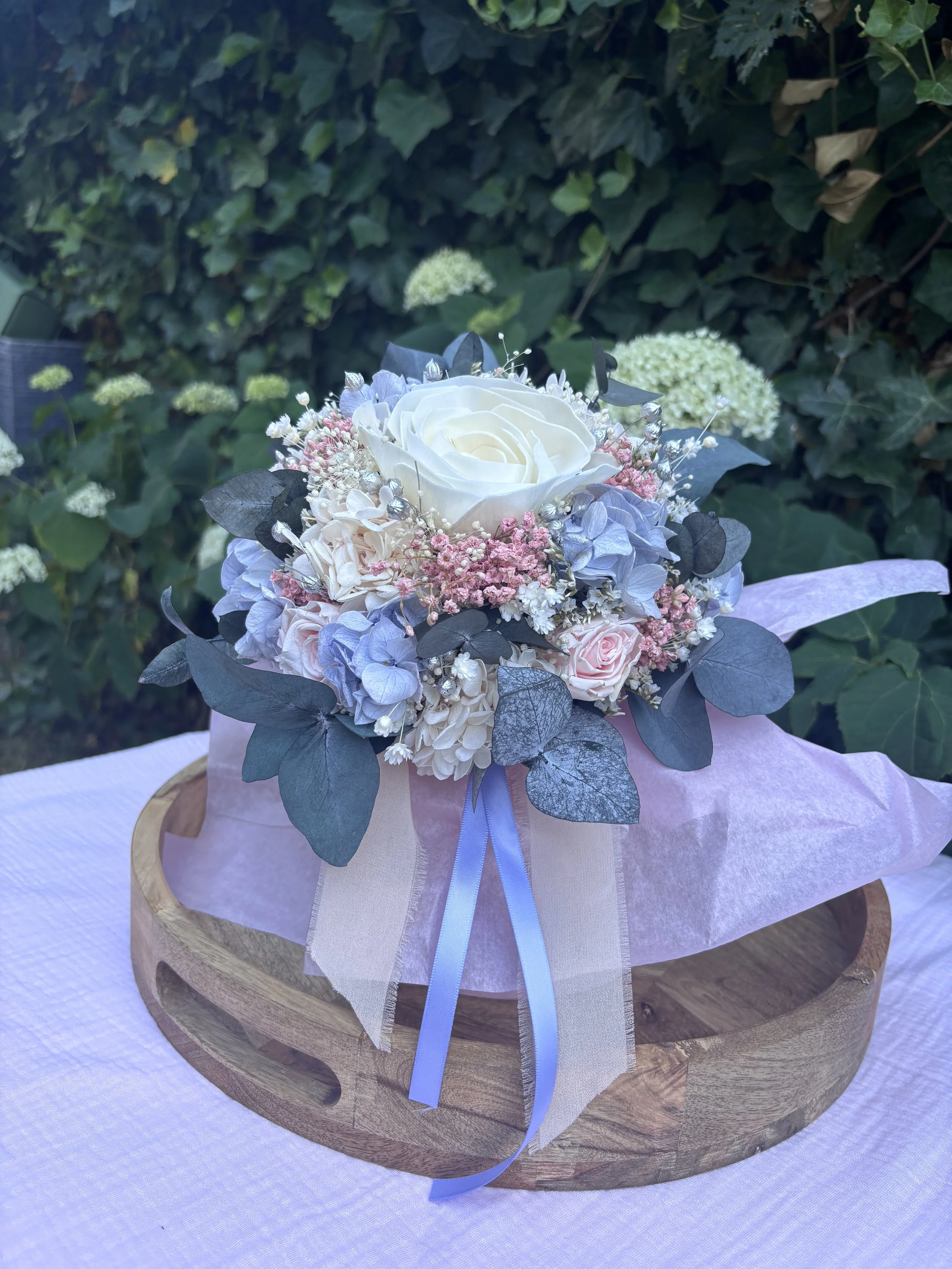 Bouquet de mariée en fleurs séchées et stabilisées aux teintes bleu pastel, rose poudré et ivoire, composé d’un camélia blanc en bois de Sola, d’hortensias stabilisés, de gypsophile, d’eucalyptus bleuté et de touches argentées. Une création romantiqu