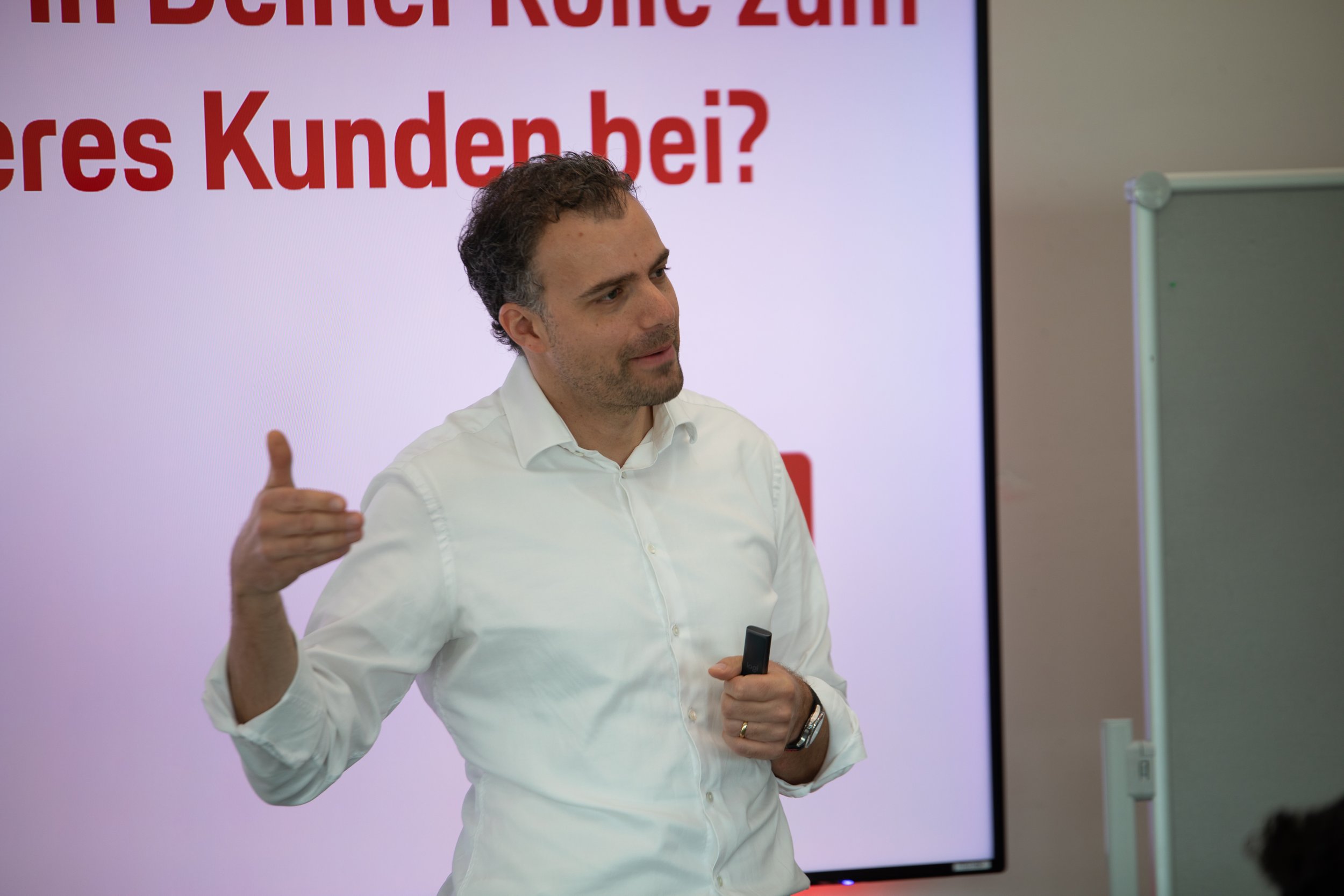 Ein Mann mit dunklem, lockigem Haar trägt ein weißes Hemd und hält ein Präsentationsgerät in der Hand. Er steht vor einem großen Bildschirm mit rotem Text, der teilweise sichtbar ist, und zeigt eine gestaltende Geste.