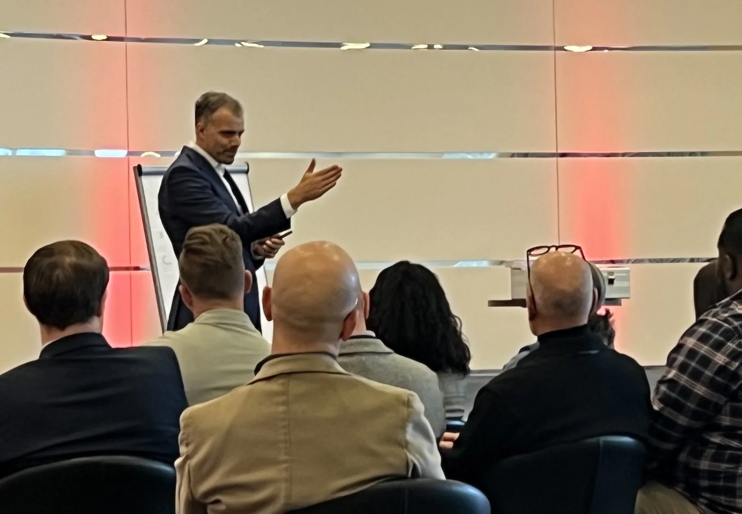 Ein Mann in einem dunklen Anzug hält eine Präsentation vor einer Gruppe von Menschen in einem Konferenzraum