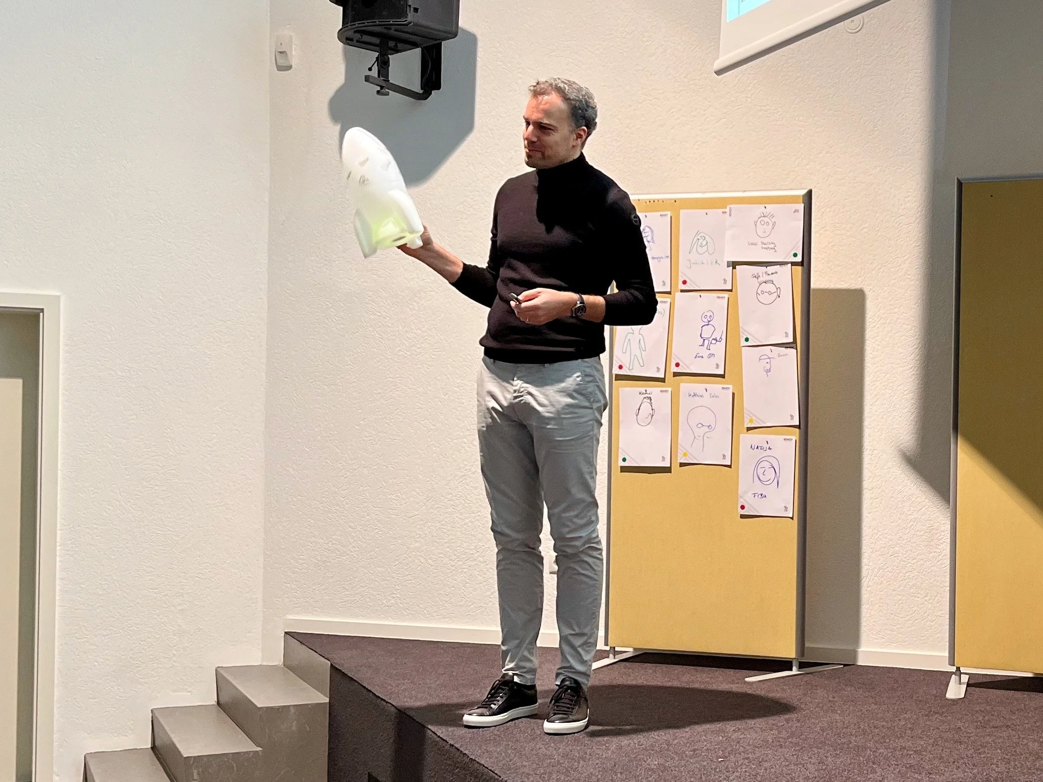 Mann steht auf einer kleinen Bühne, hält eine weiße Figur und spricht in einem Raum mit Pinnwand.