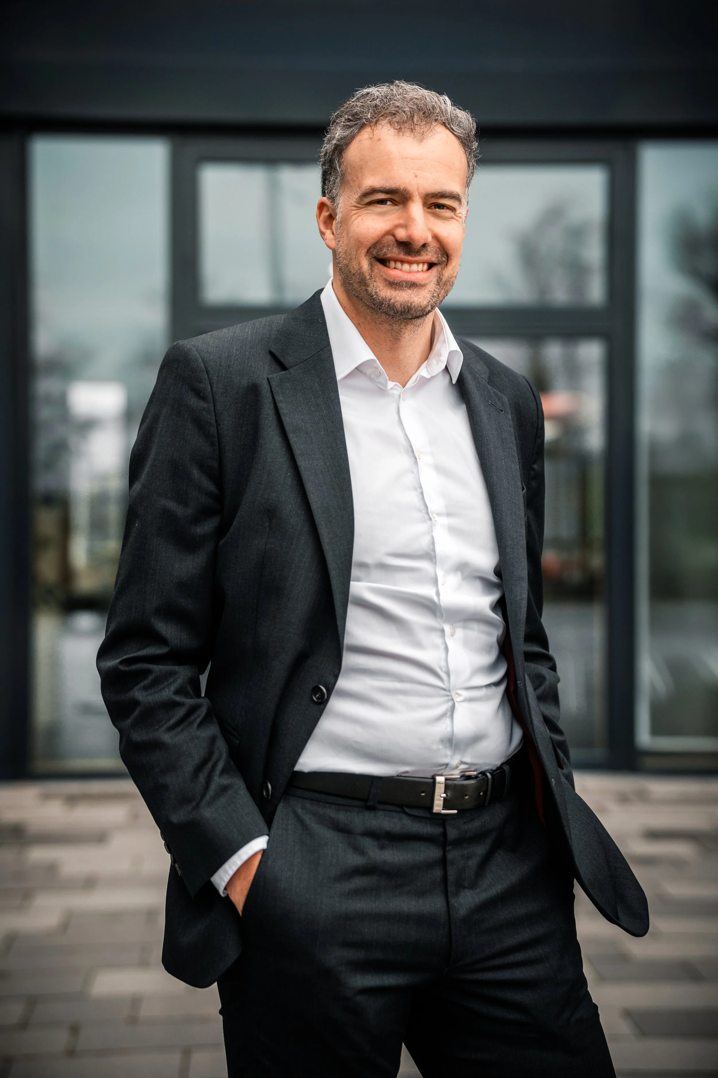 Ein Mann in dunklem Anzug und weißem Hemd steht vor einem modernen Gebäude, lächelt und hat die Hände in den Taschen.