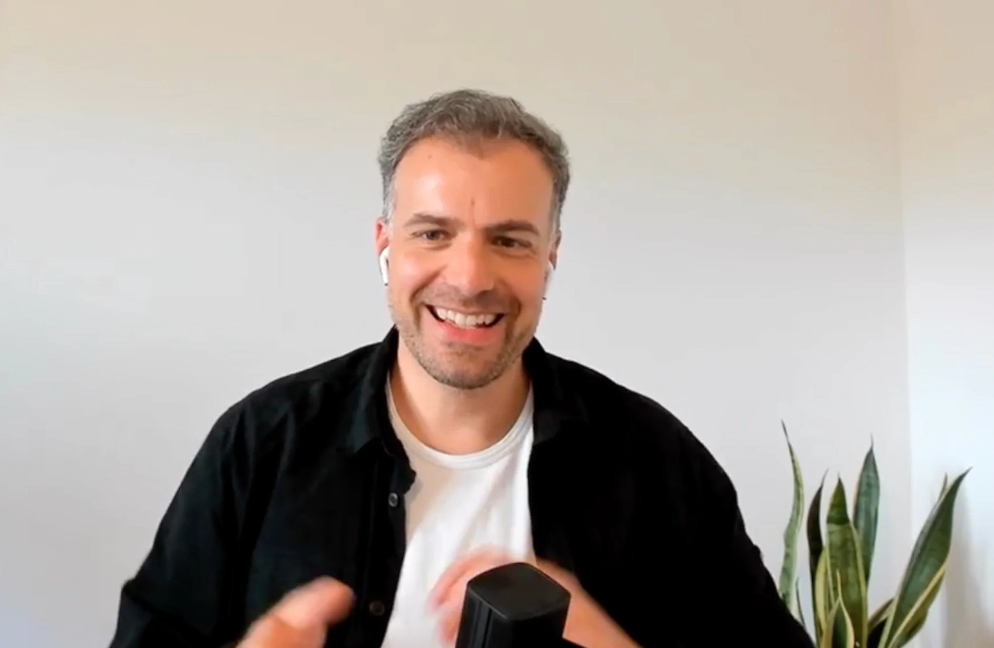 Lächelnder Mann mit grauem Haar in einem schwarzen Hemd und weißen Ohrhörern sitzt vor weißer Wand mit Pflanze im Hintergrund.