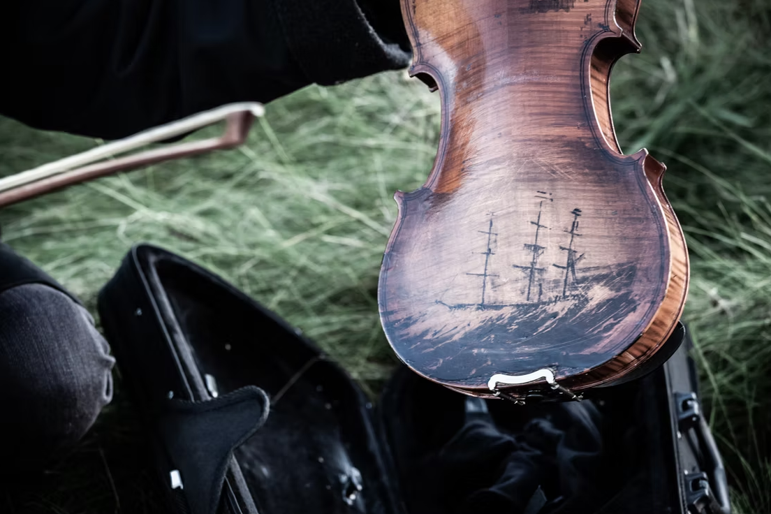 A violin with a painting of sailing ships on its back resting on grass near an open case.