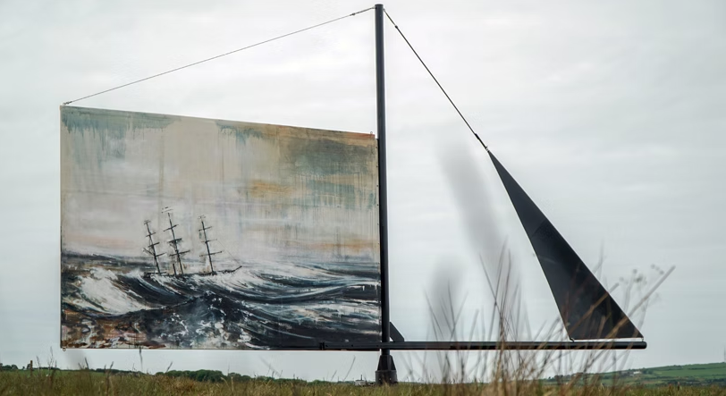 Outdoor art installation featuring a large painting of a ship at sea, mounted on a structure with a sail-like element, against a cloudy sky.
