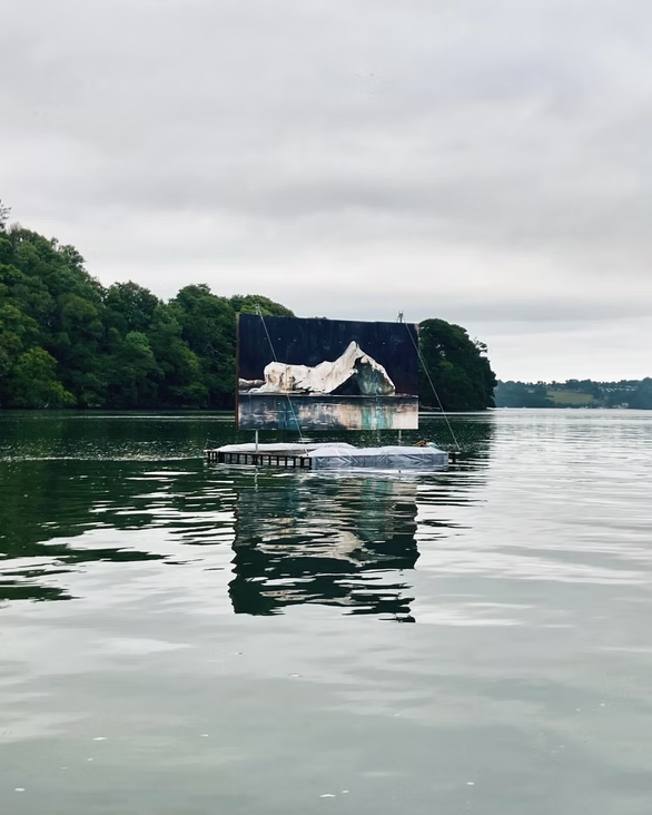 A floating platform on a lake with a large screen displaying an iceberg and distant trees under a cloudy sky.