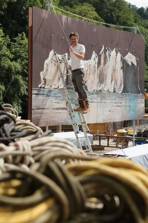 An artist stands on a ladder painting a large mural of snowy mountains near a body of water, with ropes and trees in the background.