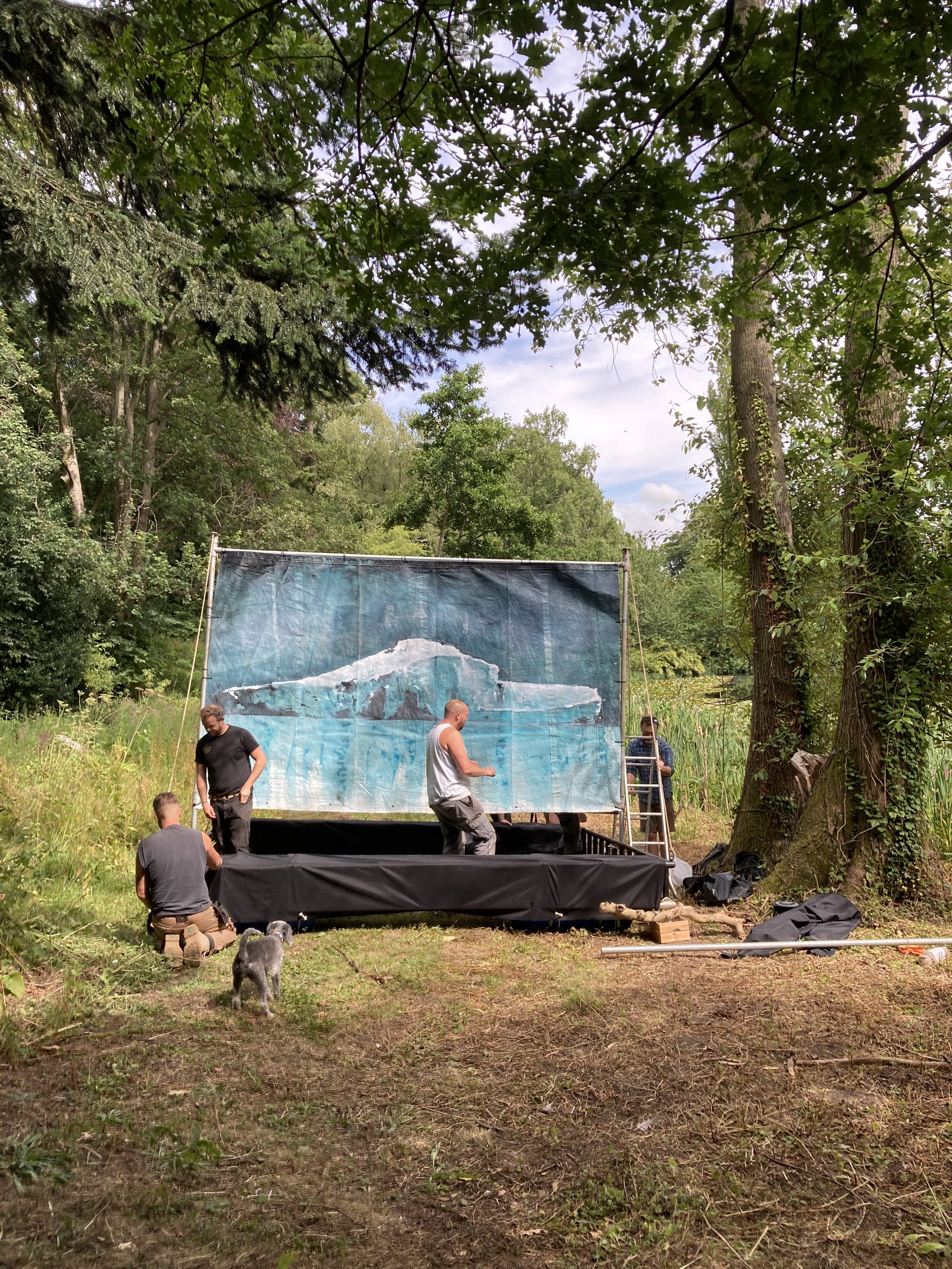 People setting up a movie screen outdoors in a wooded area with trees and grass, some of them are standing and kneeling near the screen, a dog is in the foreground.