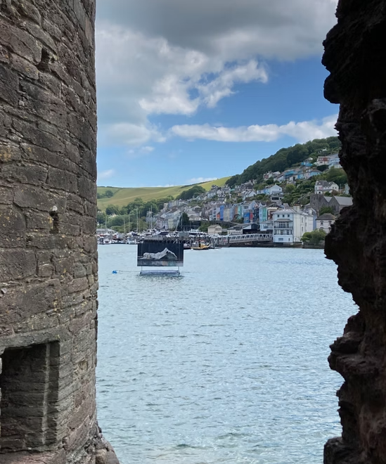 View of a harbor through a stone window, with boats on the water and houses on a hillside in the background.
