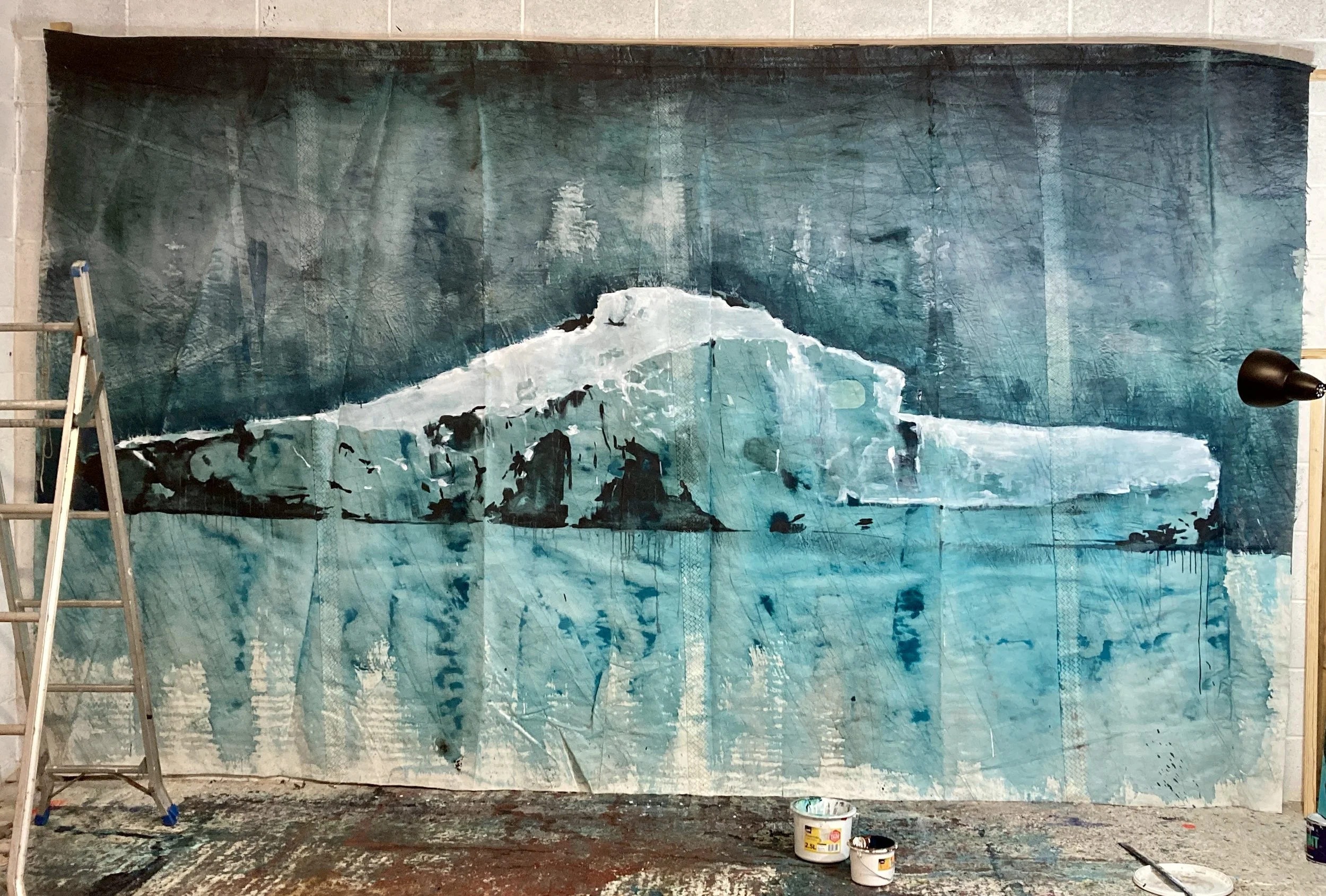 Large painting of an icy landscape, showing a glacier or iceberg in shades of white, blue, and aqua, with darker blue water and a deep blue sky in the background, on an artist's studio wall.