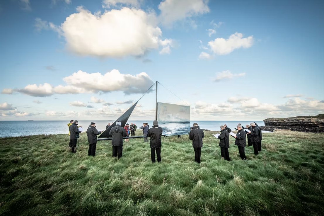 A group of people wearing dark coats and hats standing on grassy land near the ocean, observing a large reflective sculpture resembling a sailboat with a mirrored surface, against a backdrop of cloudy sky and rocky coastline.