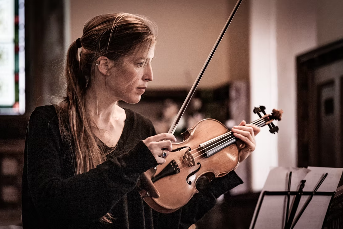 A woman with long brown hair in a ponytail, dressed in black, playing a violin with sheet music in front of her inside a warmly lit room.