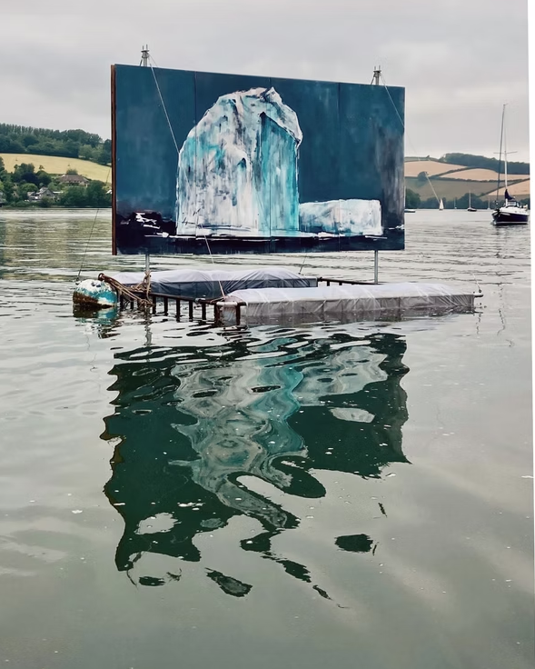 A large outdoor painting of an iceberg on a floating platform in a lake, with boats on the water and hills in the background.