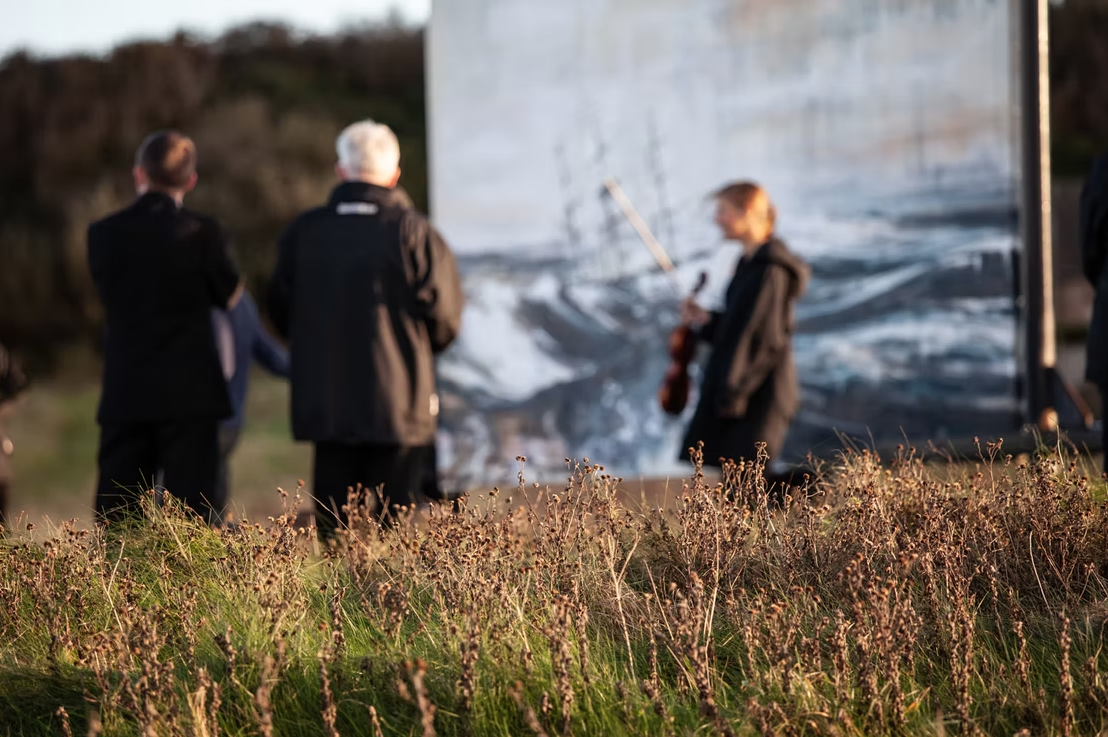 People watching a man playing guitar near a large outdoor projection screen.