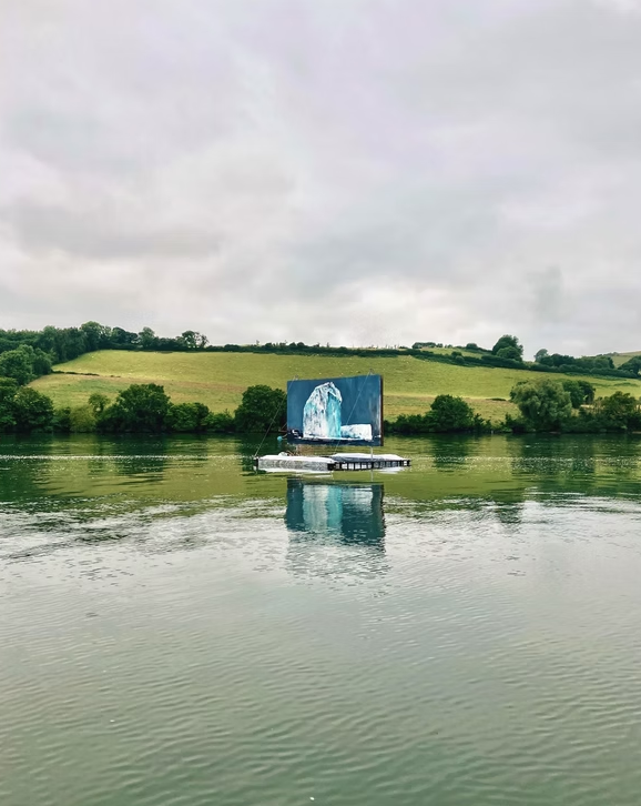 A boat with a large screen displaying a glacier image floating on a river with green hills and trees in the background under a cloudy sky.