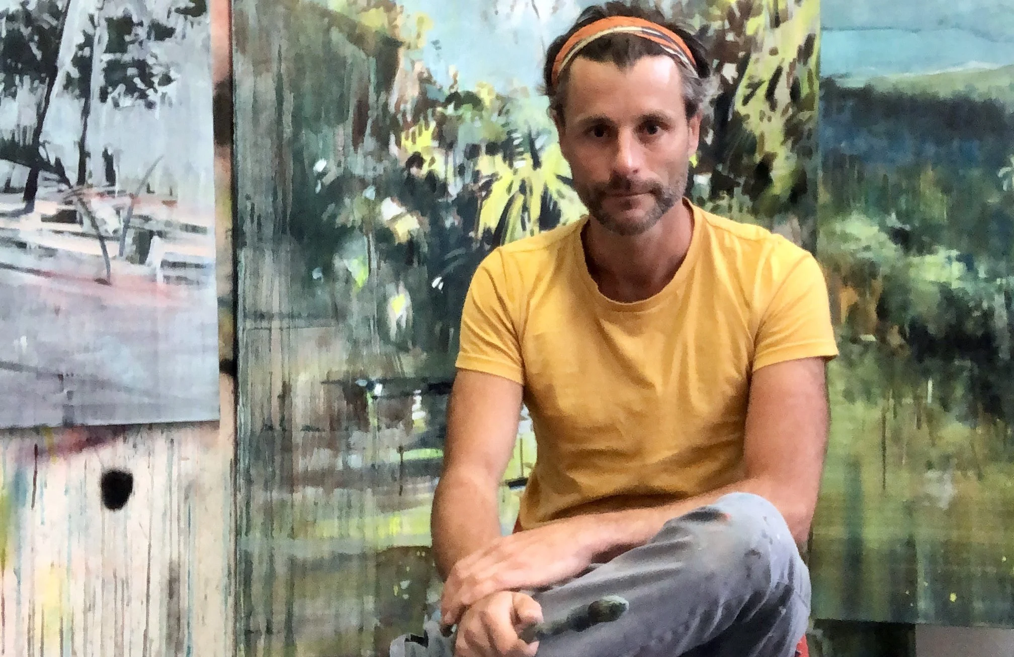 A man wearing a yellow t-shirt and gray pants sitting in front of a colorful, abstract landscape painting with trees and water.