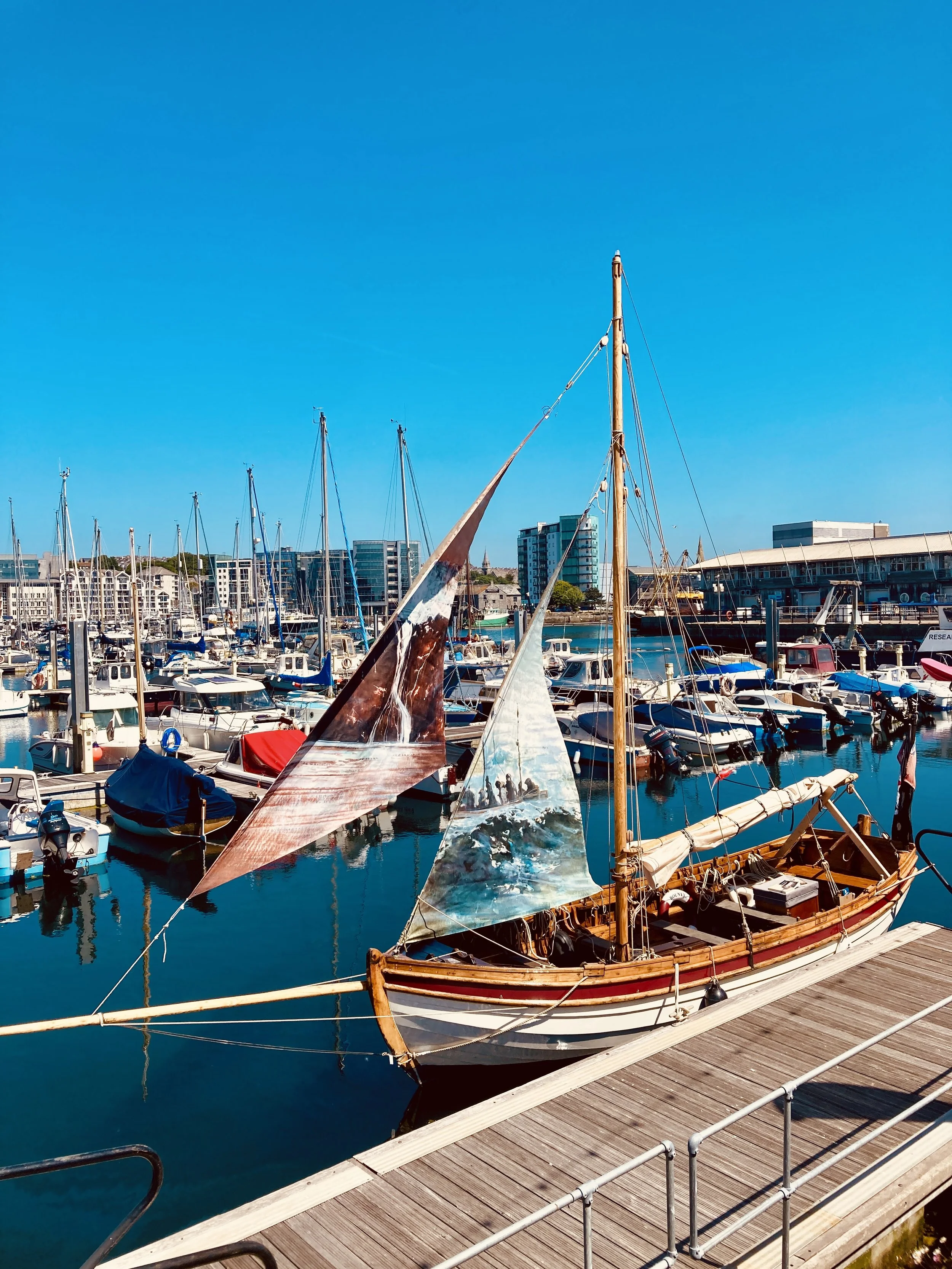 A marina with various boats and sailboats docked on calm water, with modern buildings in the background, under a clear blue sky.
