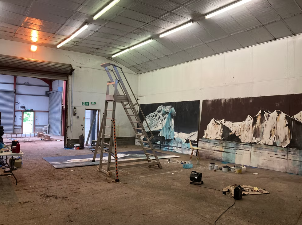 An indoor studio space with a large mural painting of icebergs on the wall. There is a ladder in the center, various painting supplies on the floor, and a chair and open door leading outside.