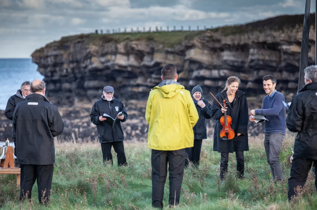 Group of people gathered outdoors near a rocky shoreline, some holding music sheets, one woman holding a violin, all engaged in a musical rehearsal.