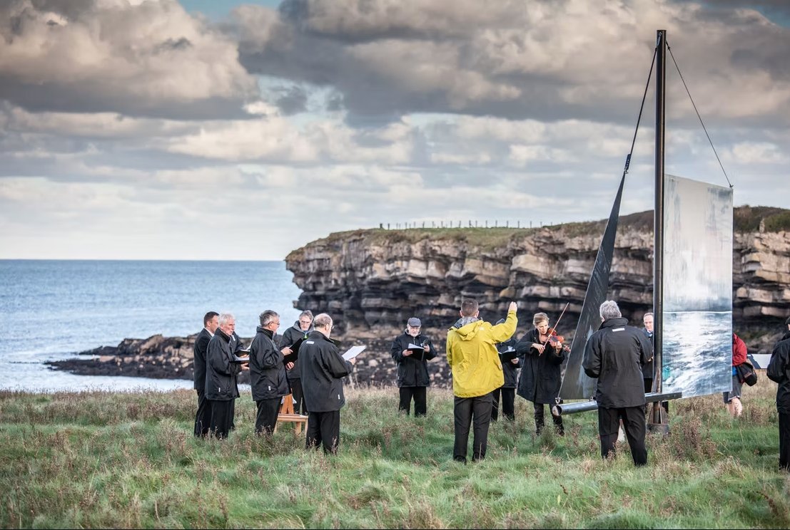 Group of people gathered outdoors near the coast, participating in a choral or musical rehearsal with a large mirror or reflective surface and a wind-swept grassy field, with cliffs and ocean in the background under cloudy sky.