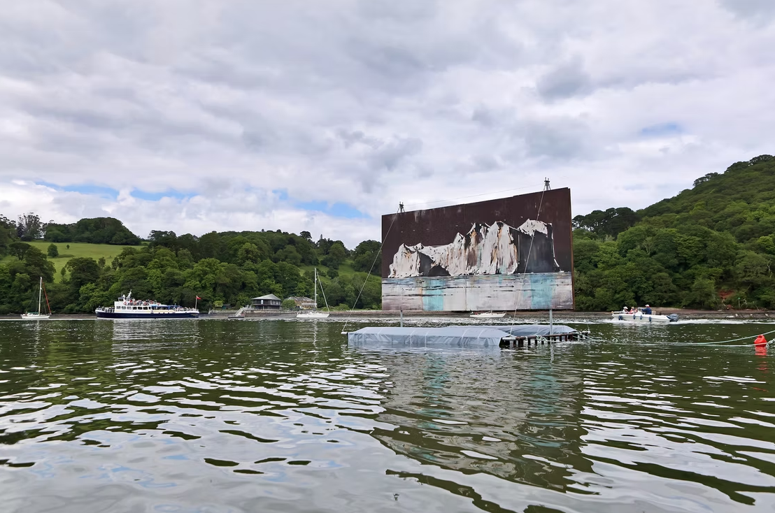A floating stage with a large screen shows an image of white cliffs with dark caves, set on a body of water with boats in the background. The surrounding landscape features lush green hills and a cloudy sky.
