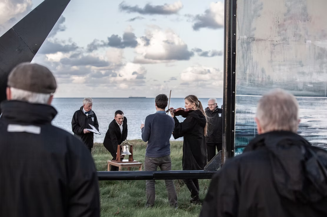 People gathered outdoors near the water with a cloudy sky in the background, participating in a ceremonial event involving a bell and a violin.