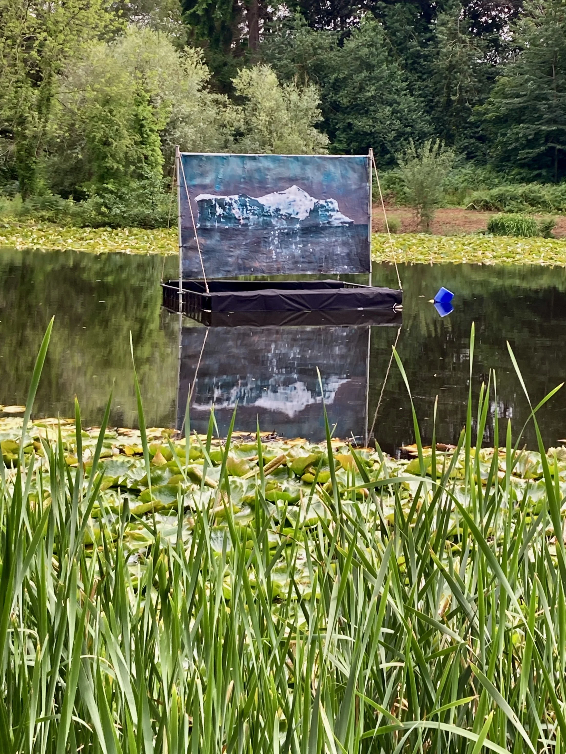 A floating art installation with a printed image of an iceberg on a fabric screen, set on a calm pond surrounded by green vegetation and trees.
