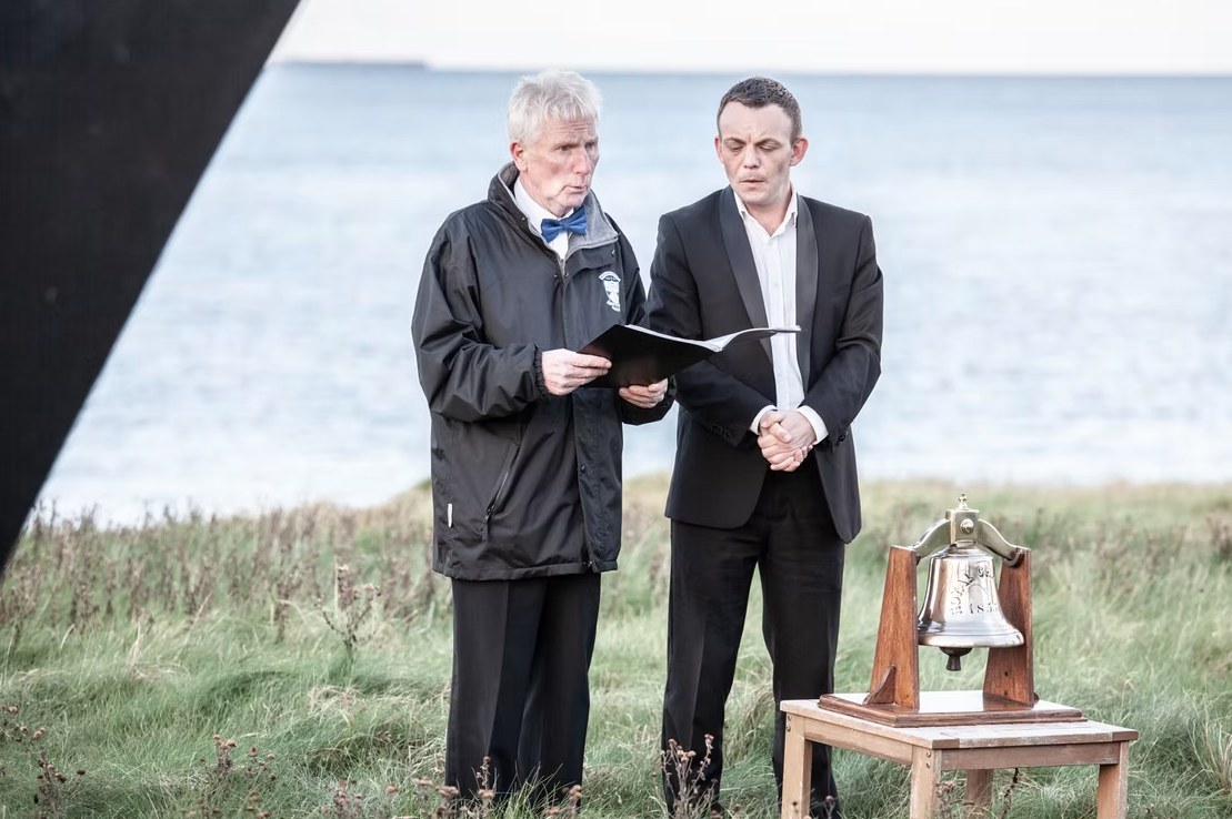 Two men standing outdoors near water, one older with gray hair, wearing a black jacket, holding a booklet, the other younger in a black suit, holding his hands together. There is a brass bell on a wooden platform nearby.