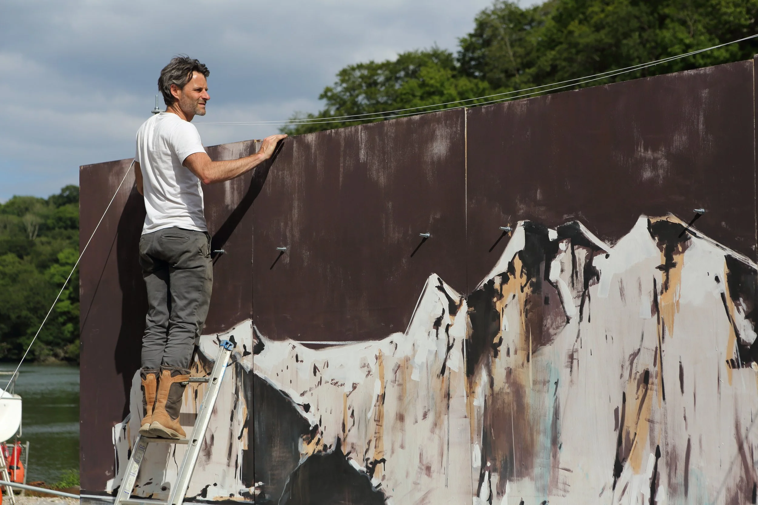 A man standing on a ladder painting a landscape on a large outdoor canvas, with trees and a river in the background.