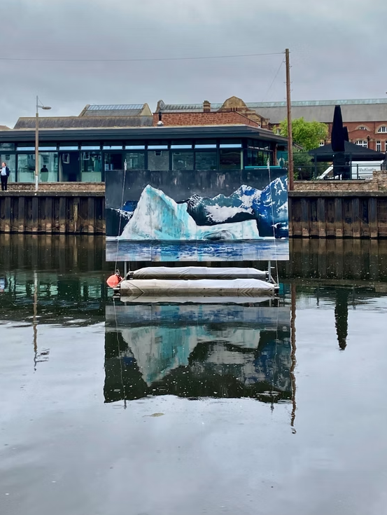 A floating art installation depicting an iceberg, situated in a body of water with a cityscape and buildings in the background.