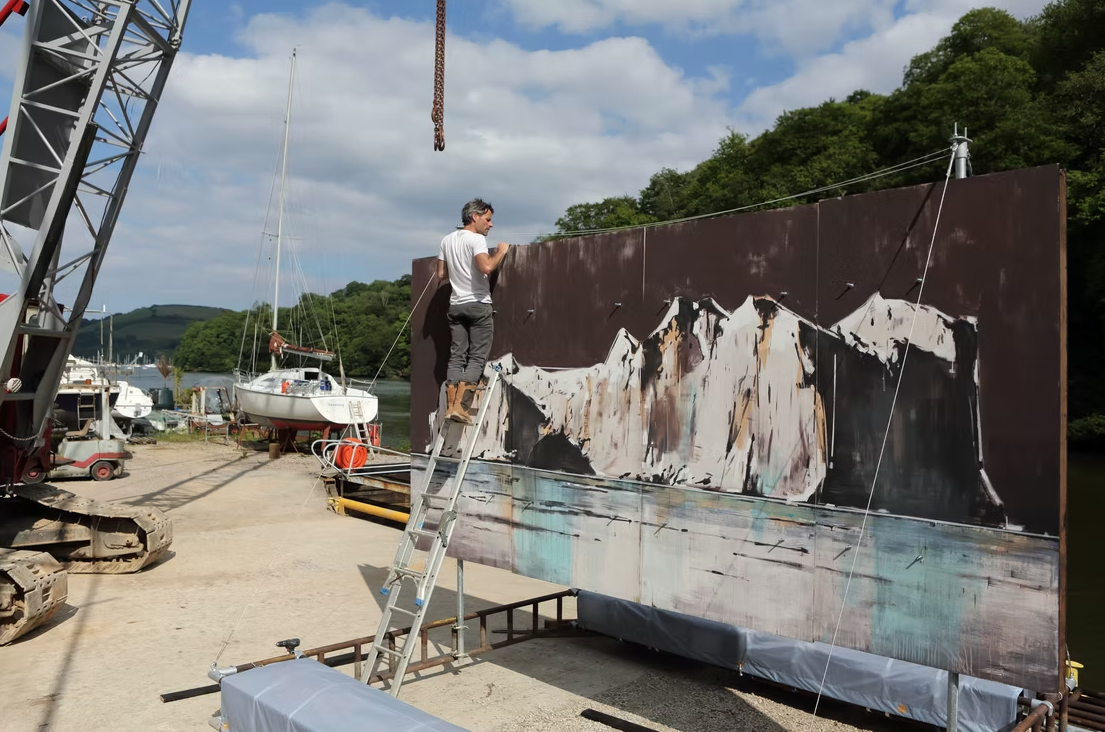 A man painting a large landscape mural of snow-capped mountains and water on a board at a marina, with boats and green hills in the background.