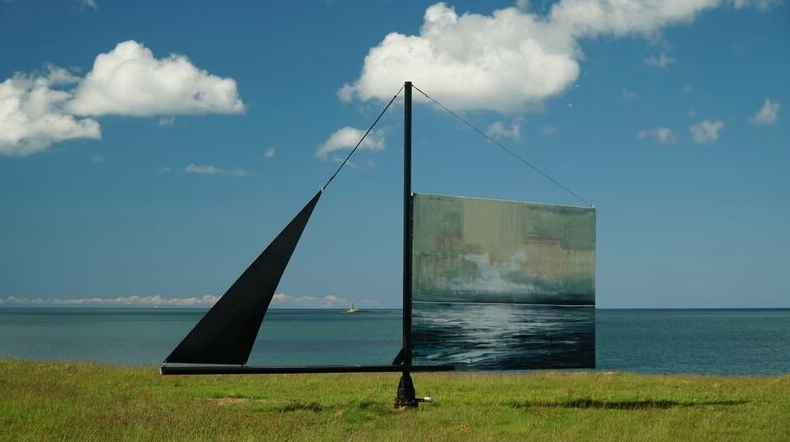Large abstract sculpture resembling a sailboat, with a tall mast and geometric shapes, located on a grassy area near the ocean under partly cloudy sky.