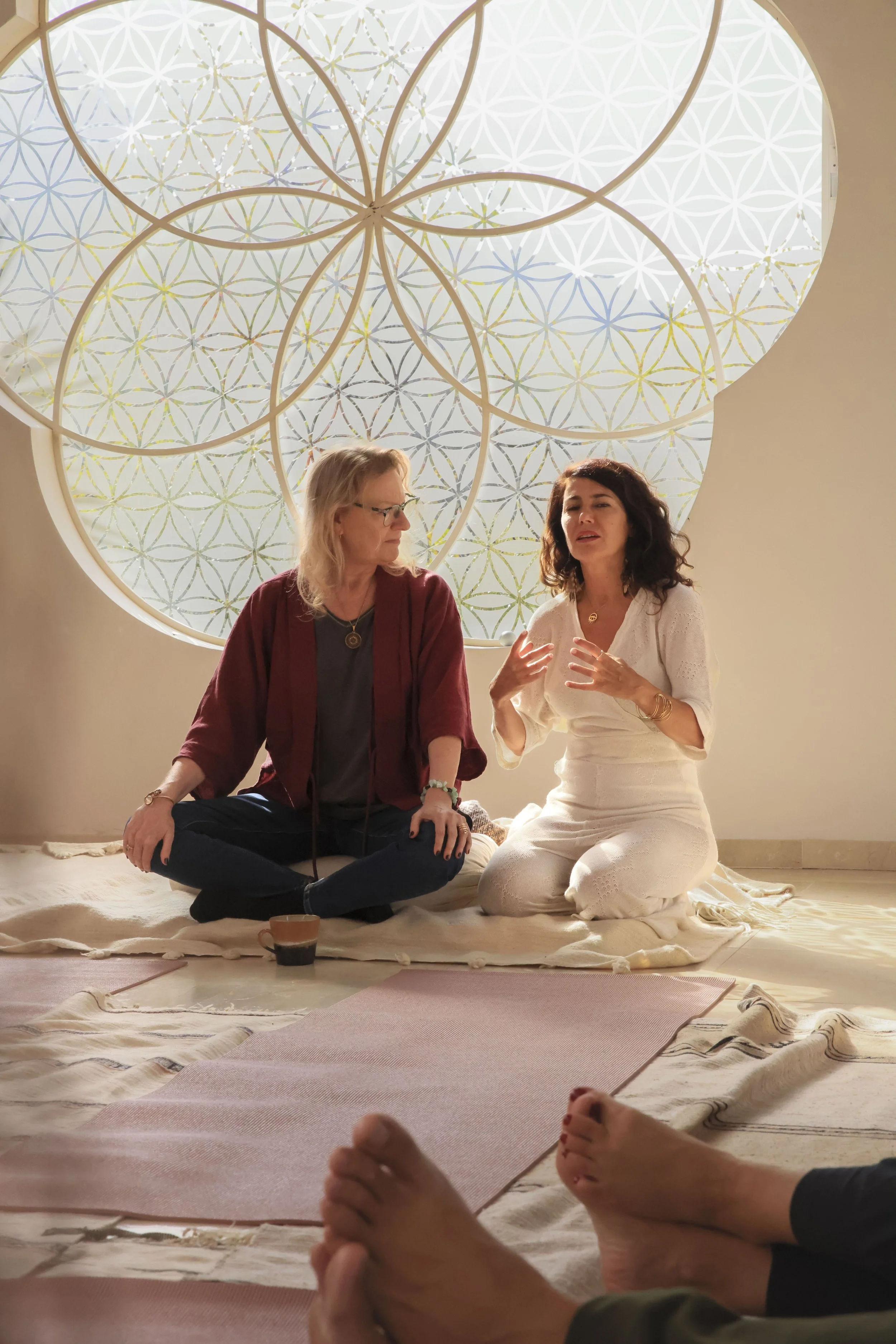 Dos mujeres sentadas en el suelo, una de ellas con un vestido blanco y la otra con una chaqueta roja, participando en una discusión o meditación, con un fondo decorativo de formas geométricas y círculos en una pared con varias alfombrillas en el suel