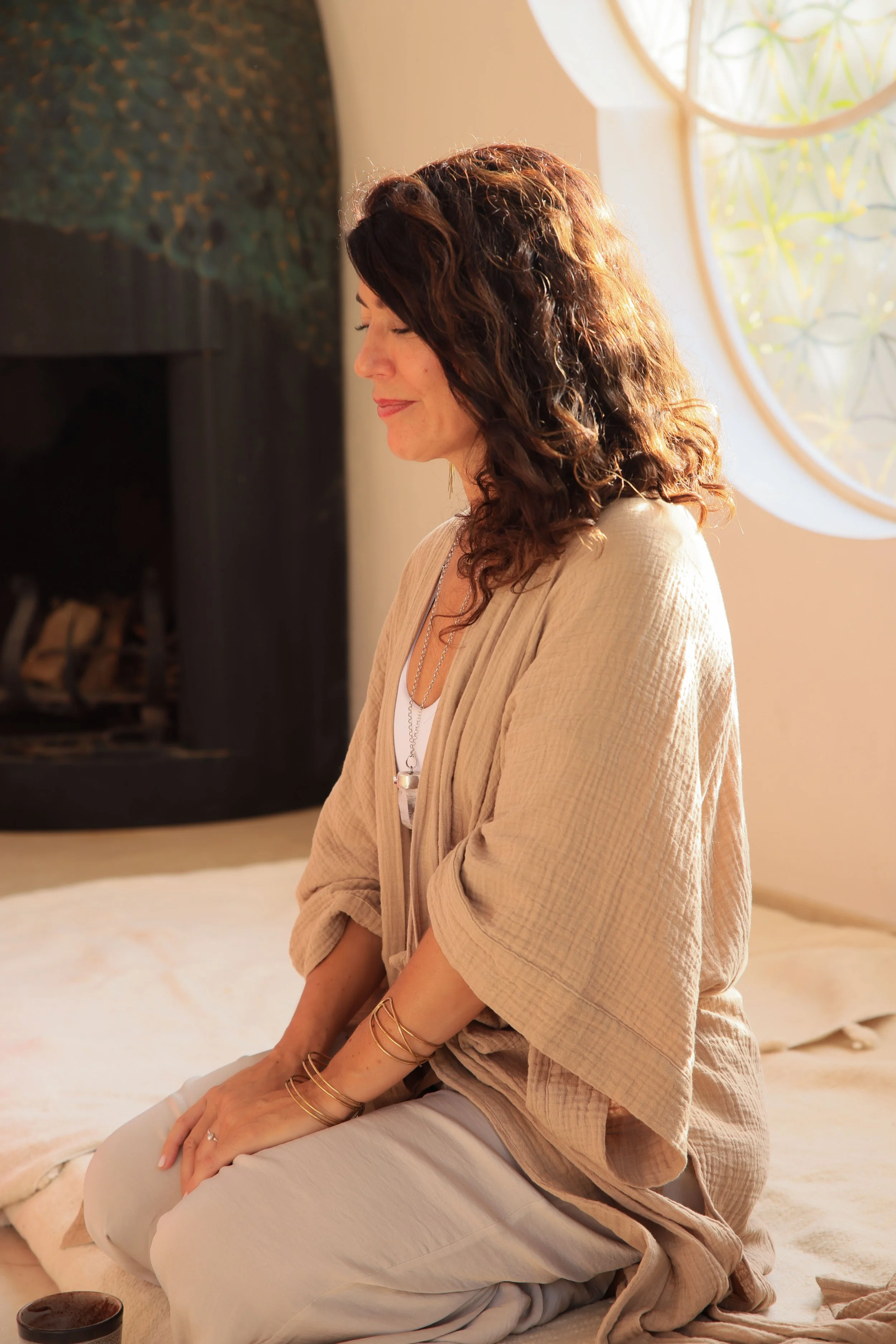 Mujer con cabello rizado, ojos cerrados, en posición de meditación en una habitación iluminada por luz natural.