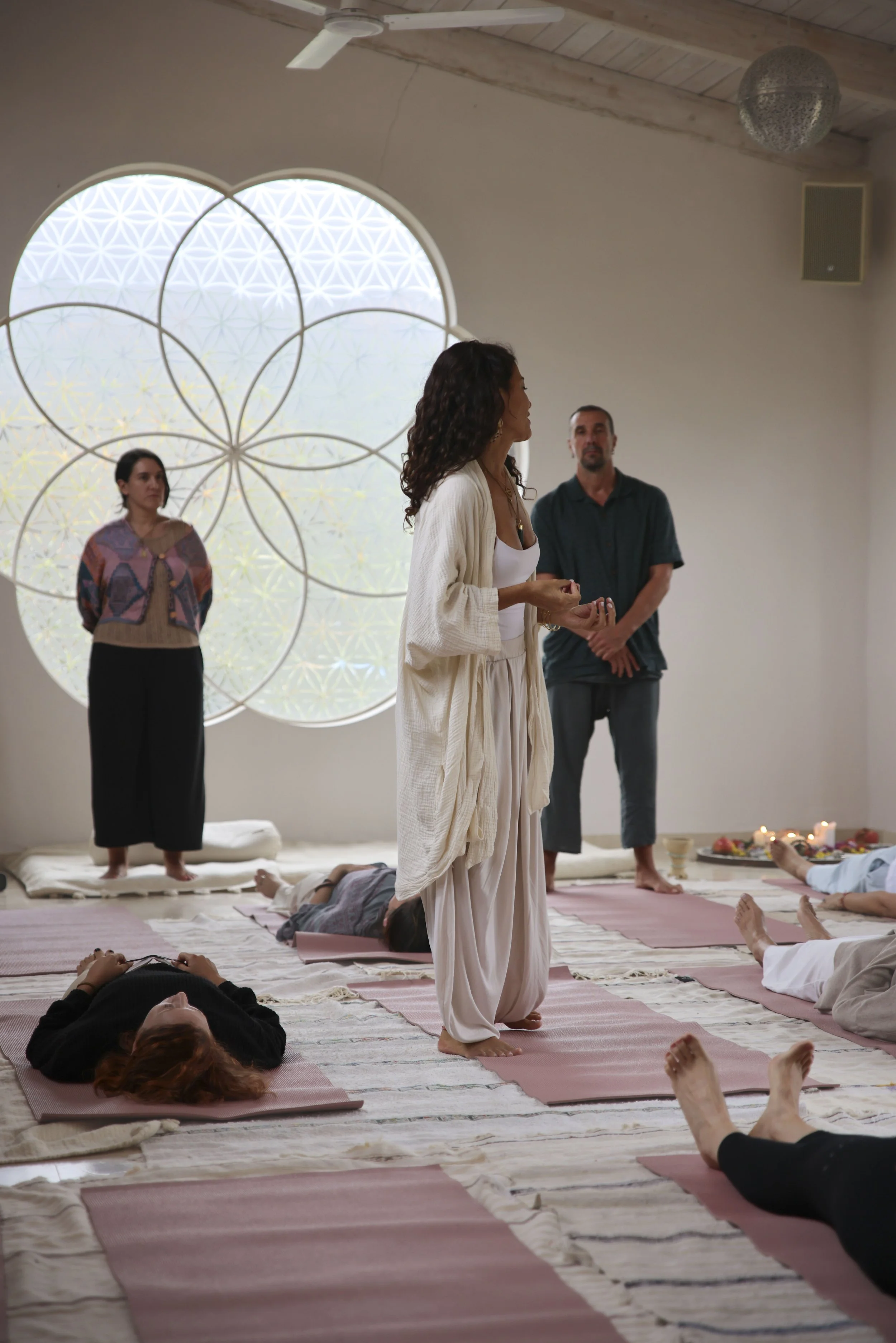 Grupo de personas participando en una clase de yoga o meditación en una sala tranquila con ventanas redondas decorativas y velas en el fondo.