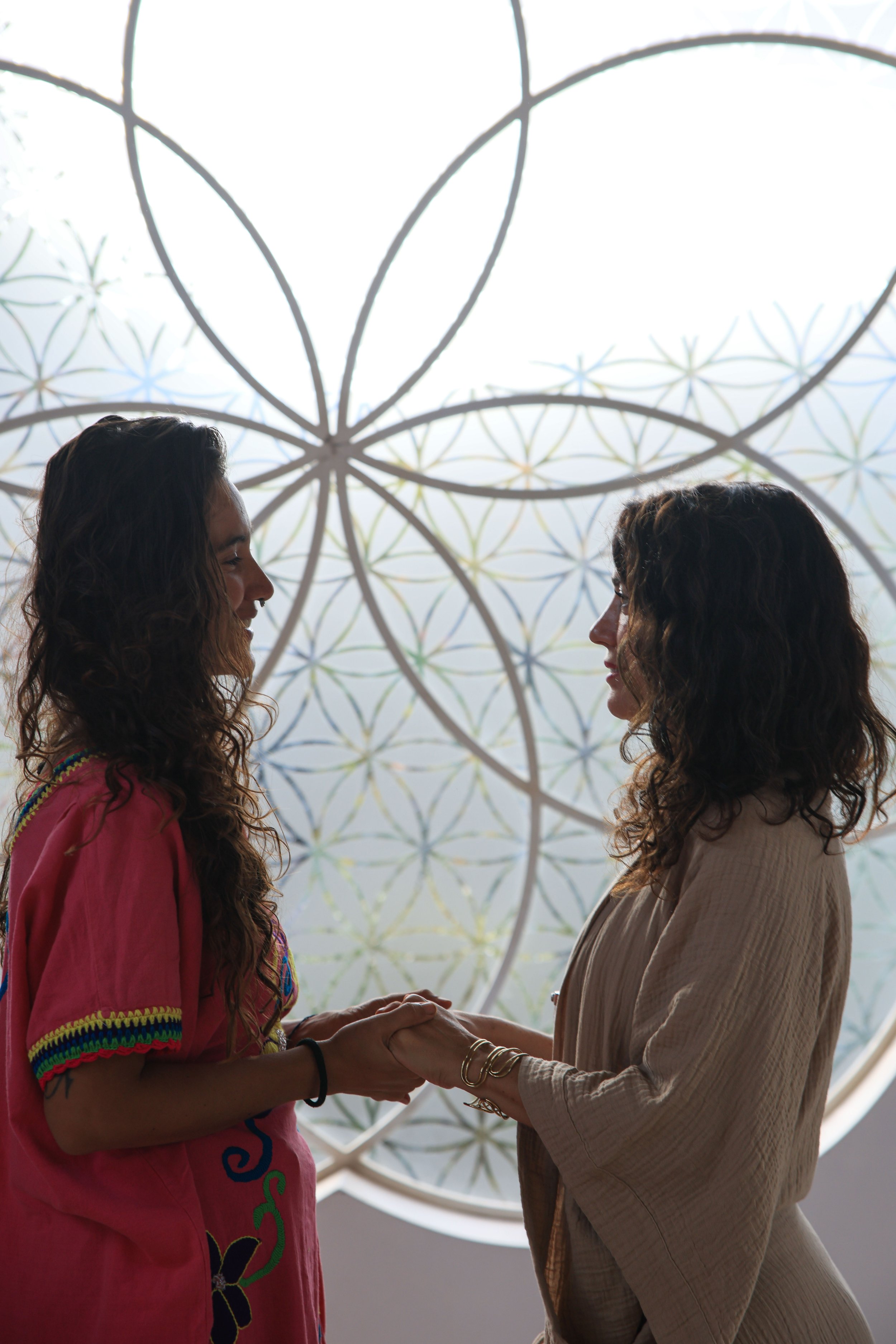 Dos mujeres entrelazando sus manos, con un fondo de vitral con patrones geométricos en un lugar interior.