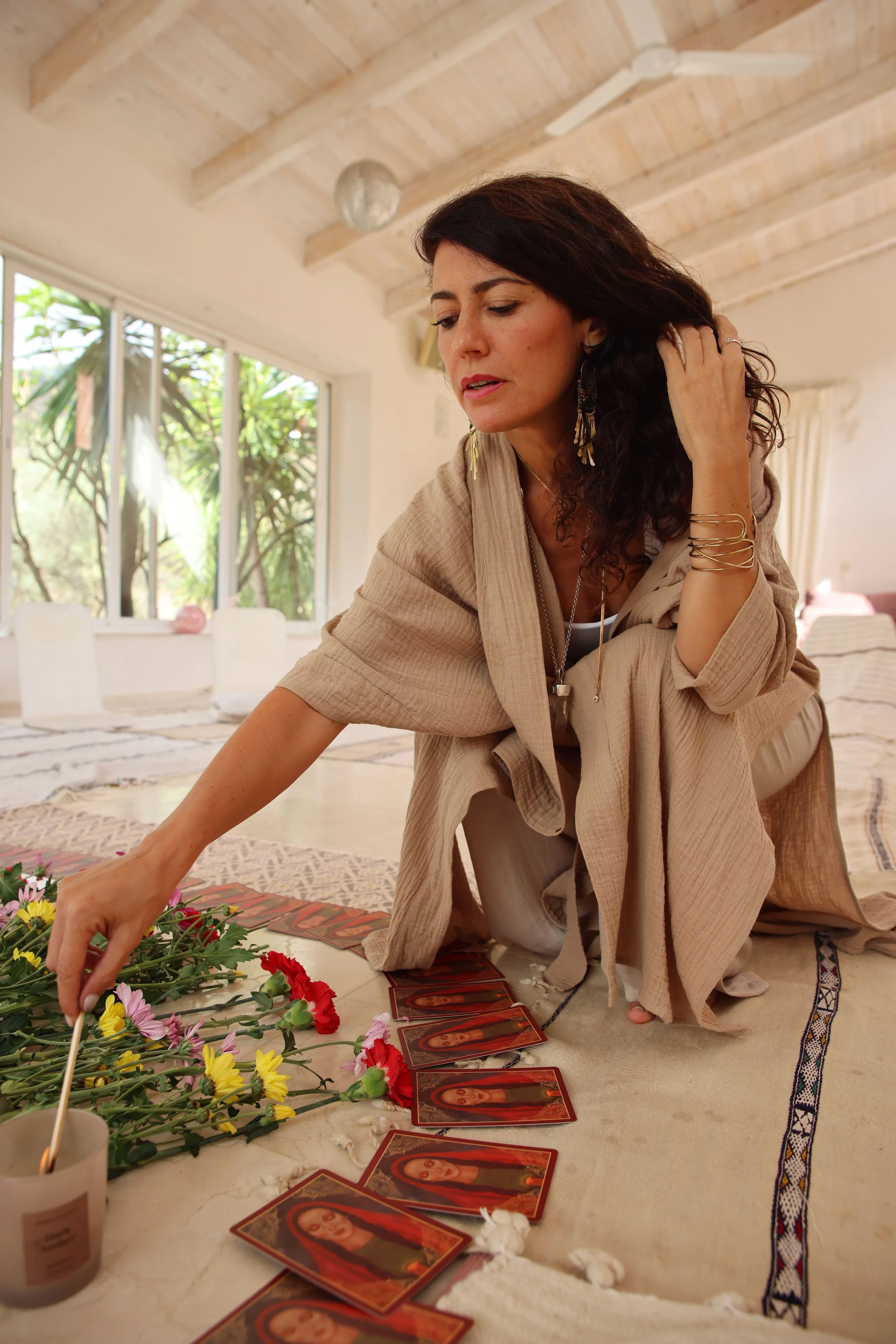 Mujer realizando una lectura de cartas de tarot en una habitación iluminada naturalmente, rodeada de flores y decoraciones tranquilas.
