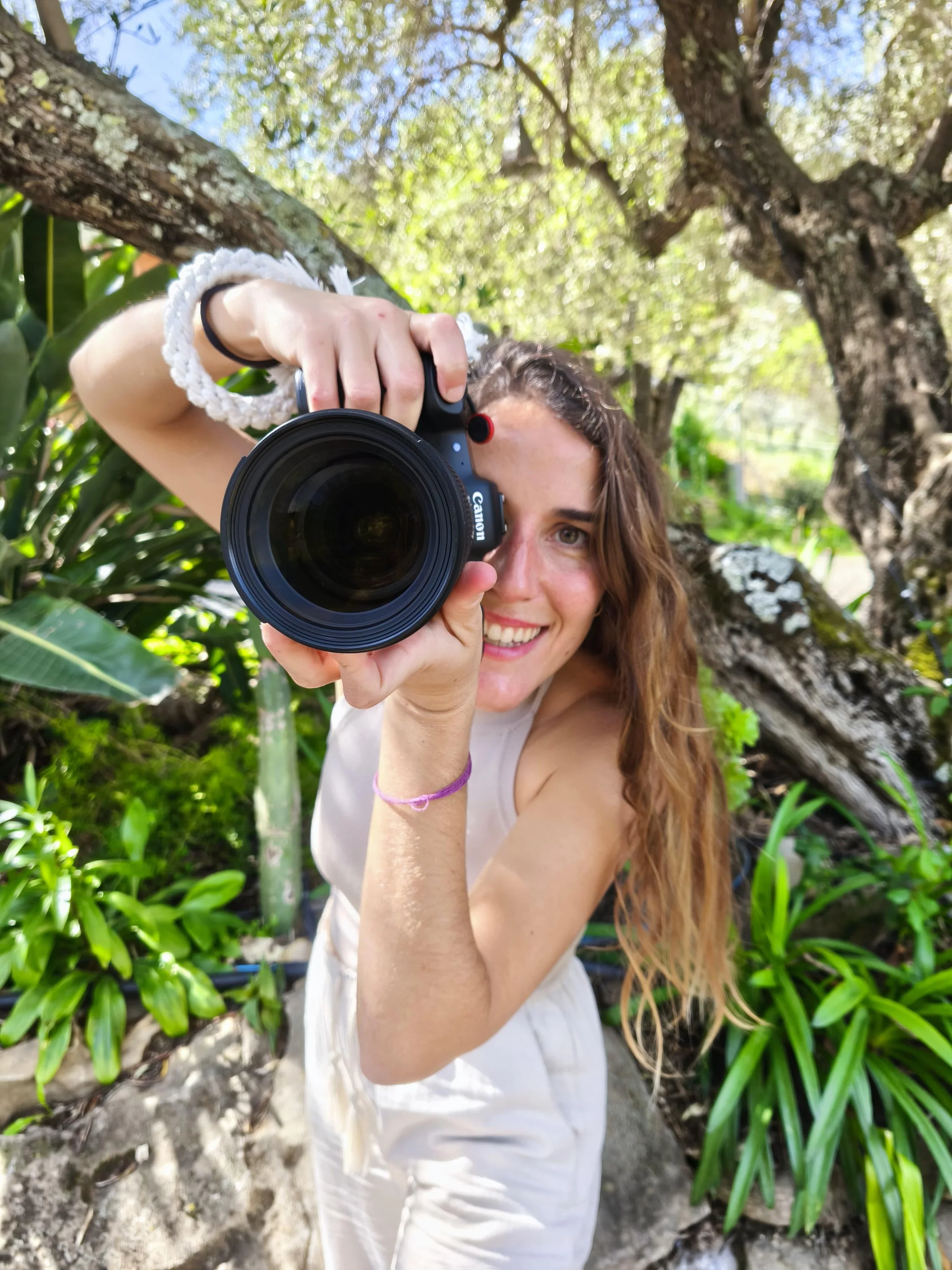 Mujer con cabello rizado y sonrisa, sosteniendo una cámara en un entorno natural con árboles y plantas verdes.