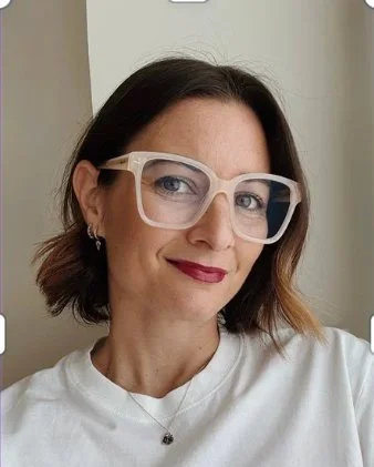 Retrato de una mujer con lentes grandes y gafas, cabello castaño y peinado en ondas, sonriendo y usando un collar sencillo, con fondo neutro.