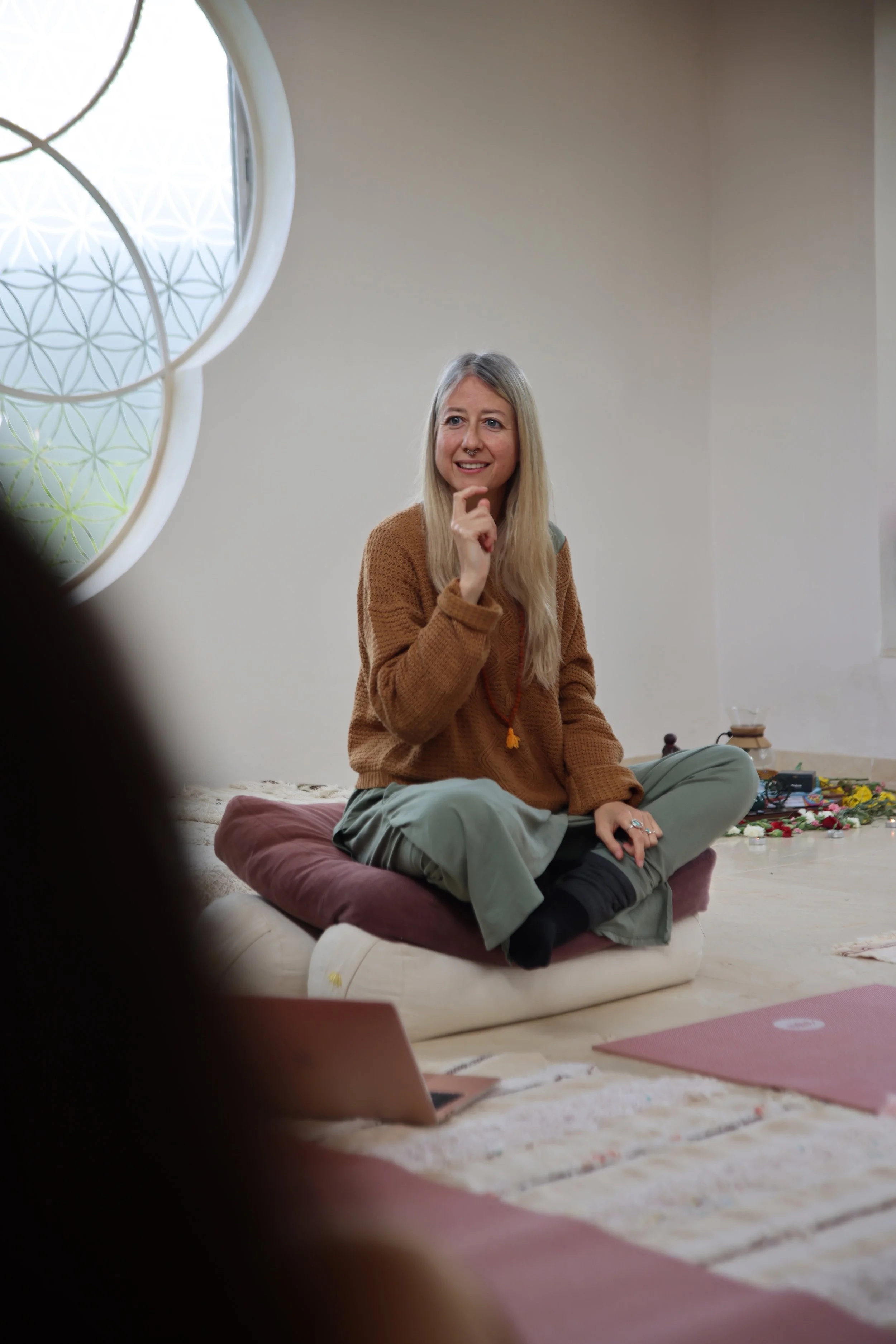 Mujer con cabello rubio y suelto, vestida con suéter marrón y pantalones verdes, sentada en un cojín en el suelo en una sala con ventanas redondas decoradas, rodeada de velas, flores y artículos de yoga, sonriendo y haciendo gesto con la mano.