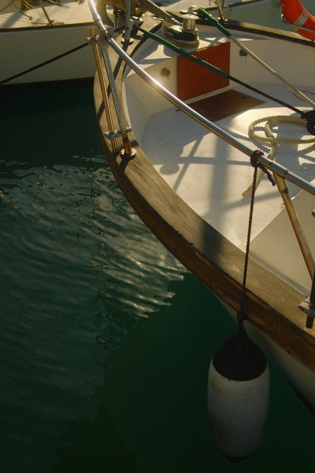 Close-up of a boat's bow with a wooden trim, metal railing, ropes, and a white buoy hanging from the side, floating on calm water.