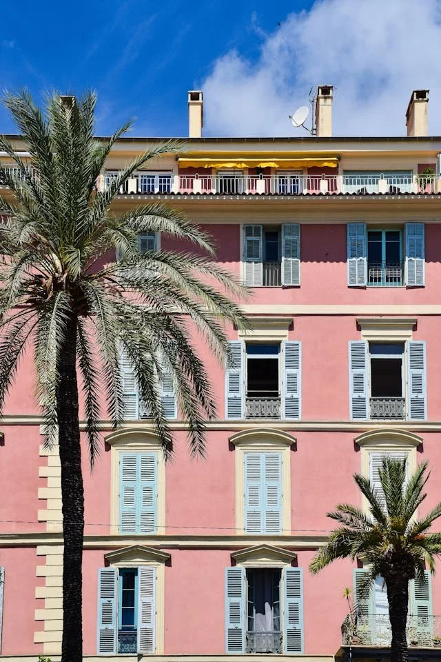 Pink European-style building with multiple windows and white shutters, two palm trees in front, bright blue sky with some clouds.