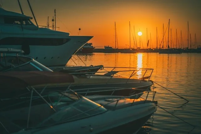 Boats docked at a marina during sunset with the sun low in the sky and reflections on the water.
