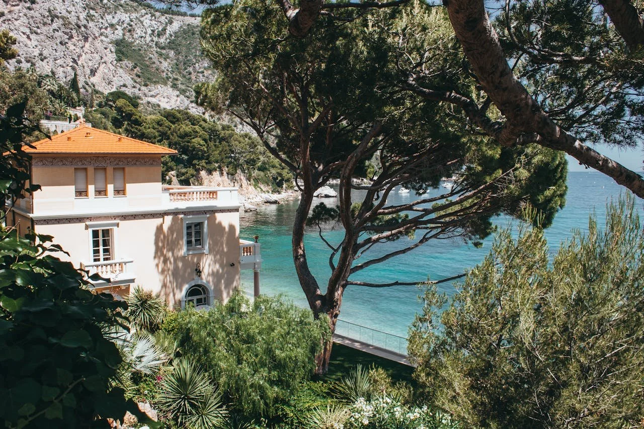 A house with a peach-colored roof is surrounded by lush green trees on a coastline with turquoise water and rocky cliffs in the background.