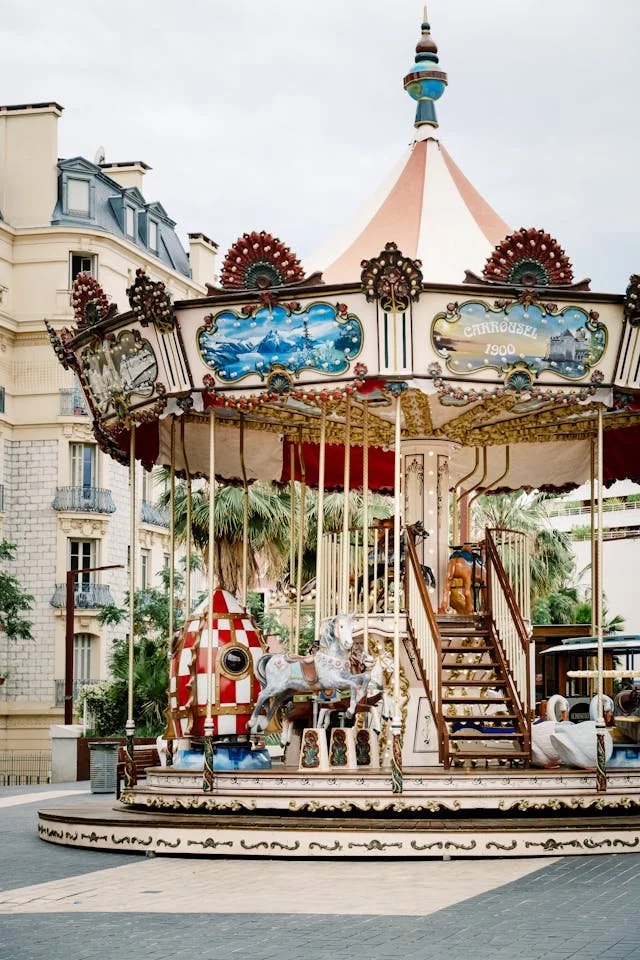 A vintage carousel with a pink and cream canopy, featuring painted scenery and ornate decorations, including a white and red rocket and a horse with a silver mane, set in an outdoor area with surrounding buildings and palm trees.