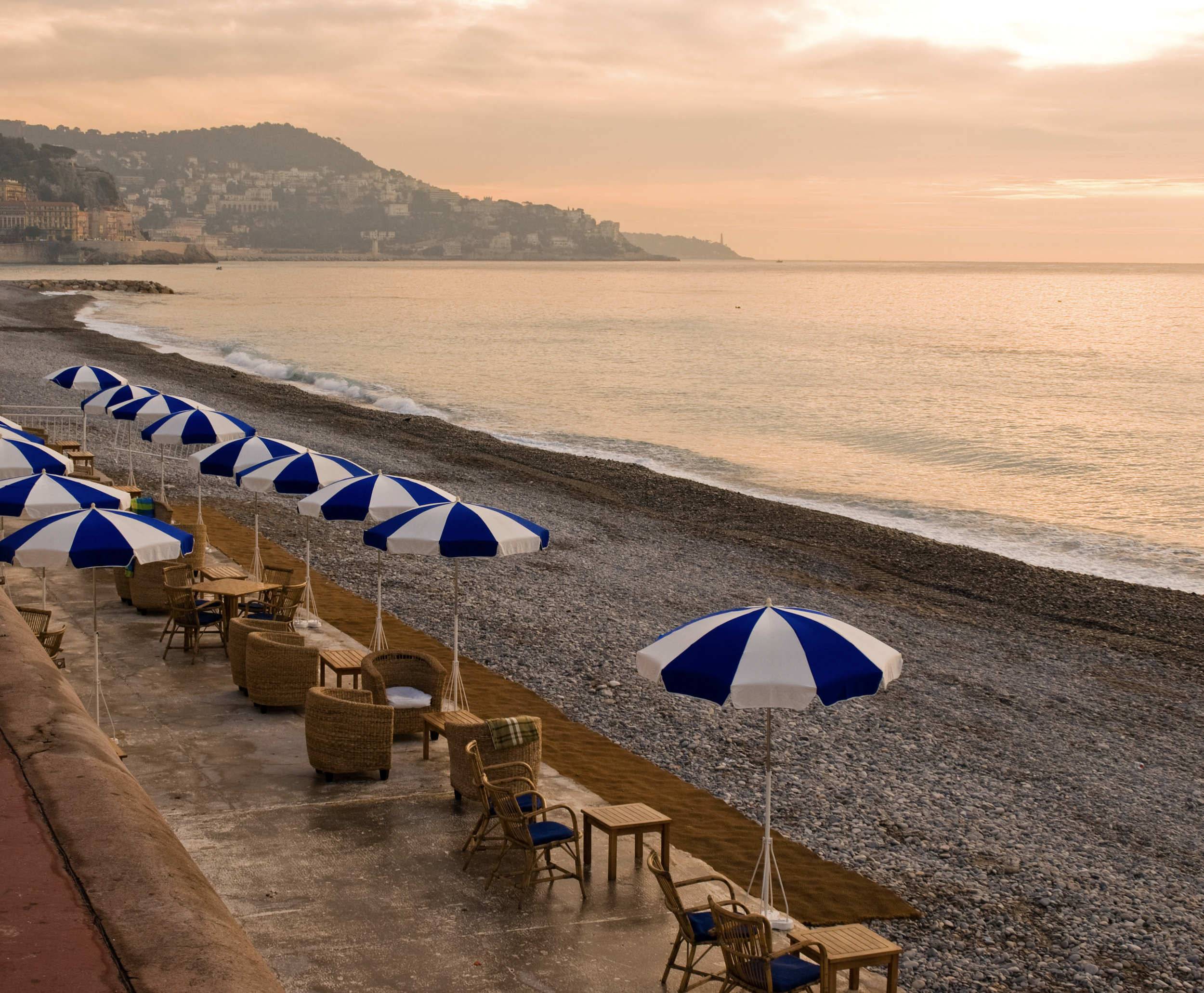 Empty beach with blue and white umbrellas, wicker chairs, and wooden tables at sunrise or sunset, overlooking calm water with distant hills.