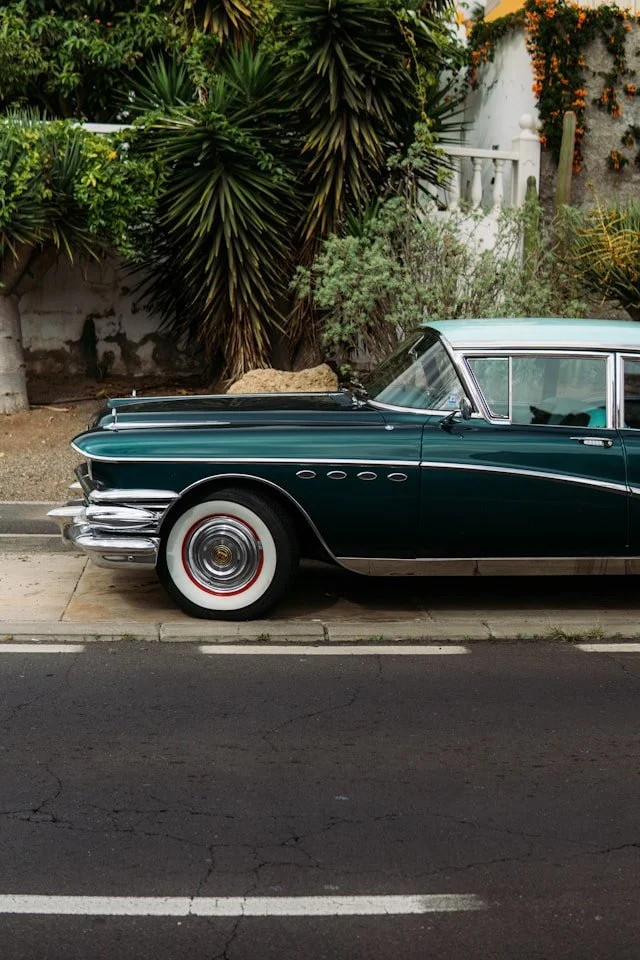 A vintage dark green car parked on a street beside a sidewalk with plants and a white fence in the background.