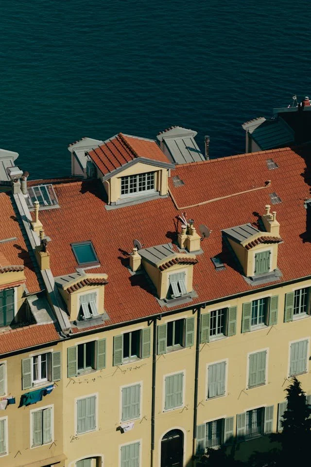 A view of a yellow building with green shutters and a red tiled roof near the water, with several dormer windows and rooftop features.